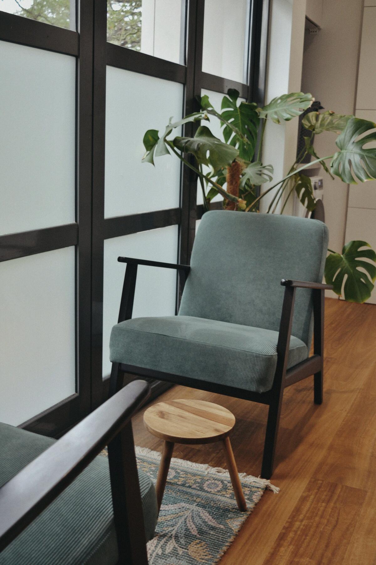A green armchair and a small wooden table beside tall windows and plants