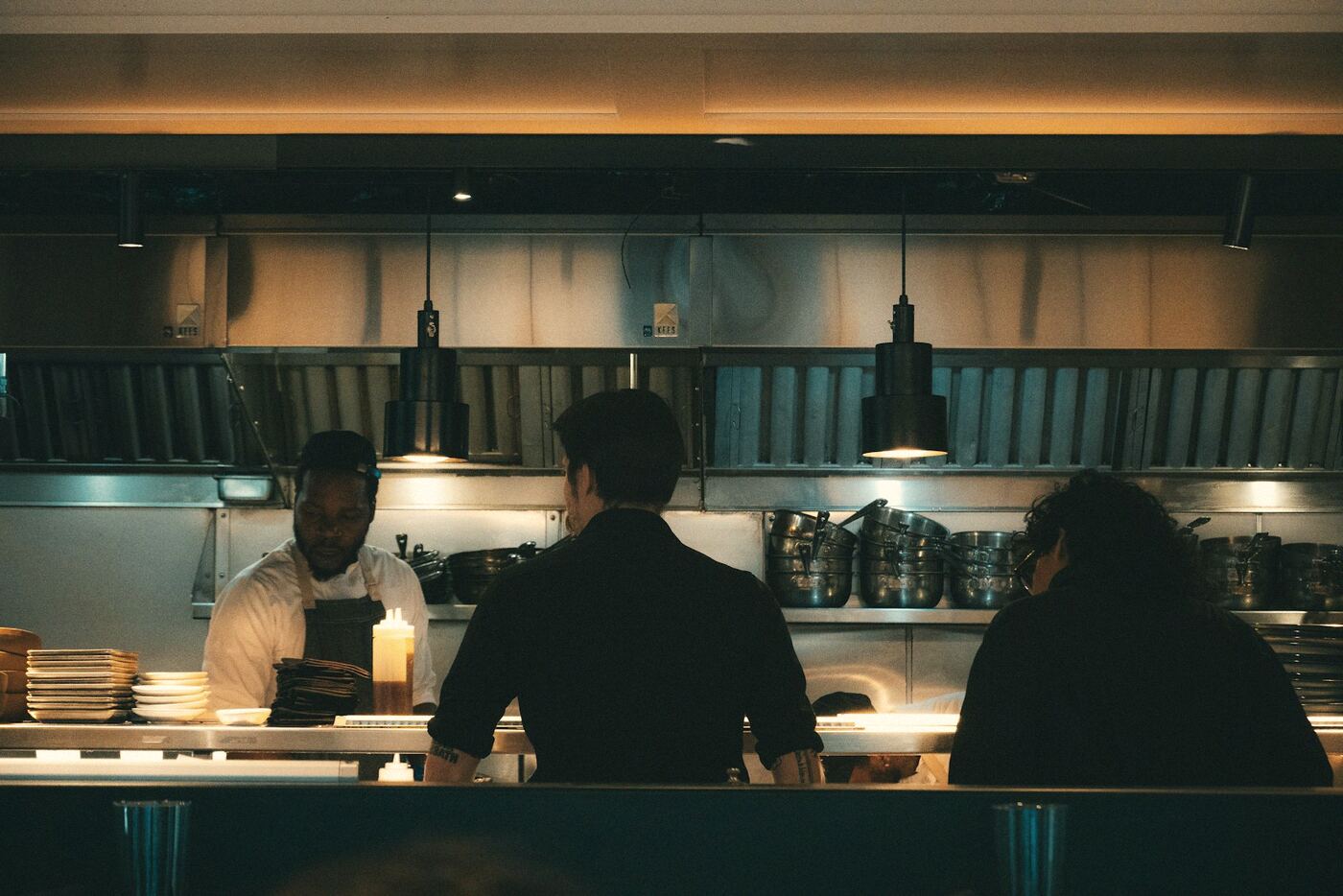 Cooks at the pass of a dim open kitchen under pendant lamps